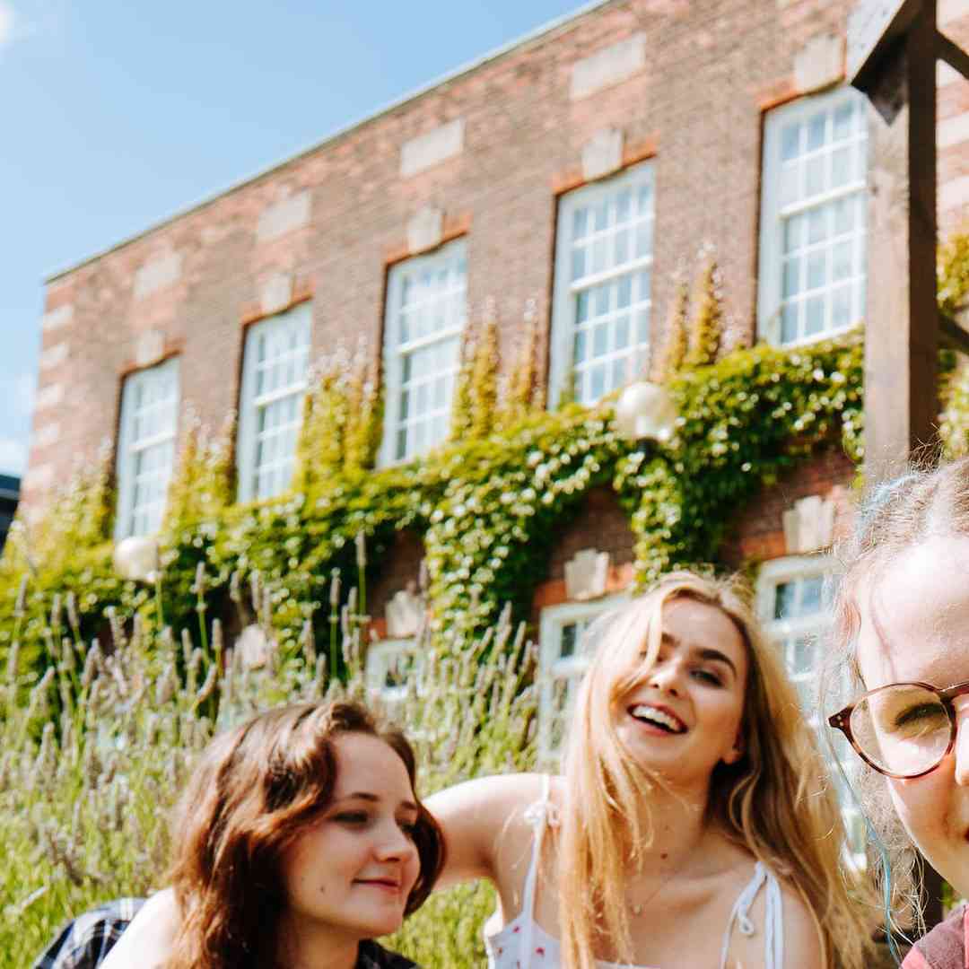 A group of students smile and laugh outside a red-brick, ivy-clad University building. 