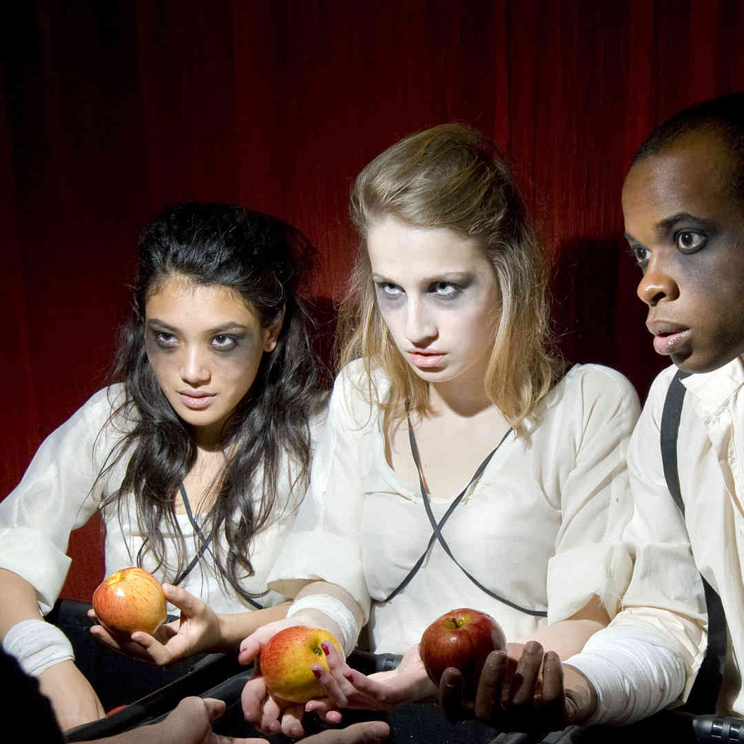 Three students performing a play called 'Our Little Secret'. They're wearing dark eye makeup and are handing out apples.