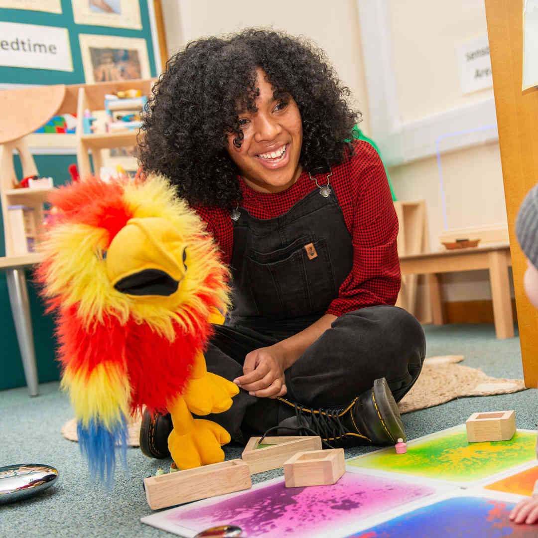 A teacher smiles at a baby while playing with a parrot puppet in a classroom