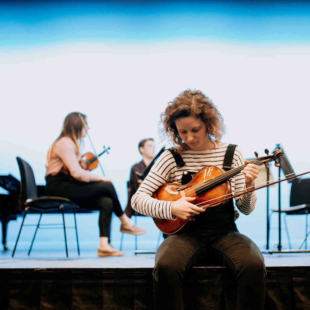 Music student, Anna Kent, sits on the edge of a stage tuning a violin in front of the rest of their quartet members.