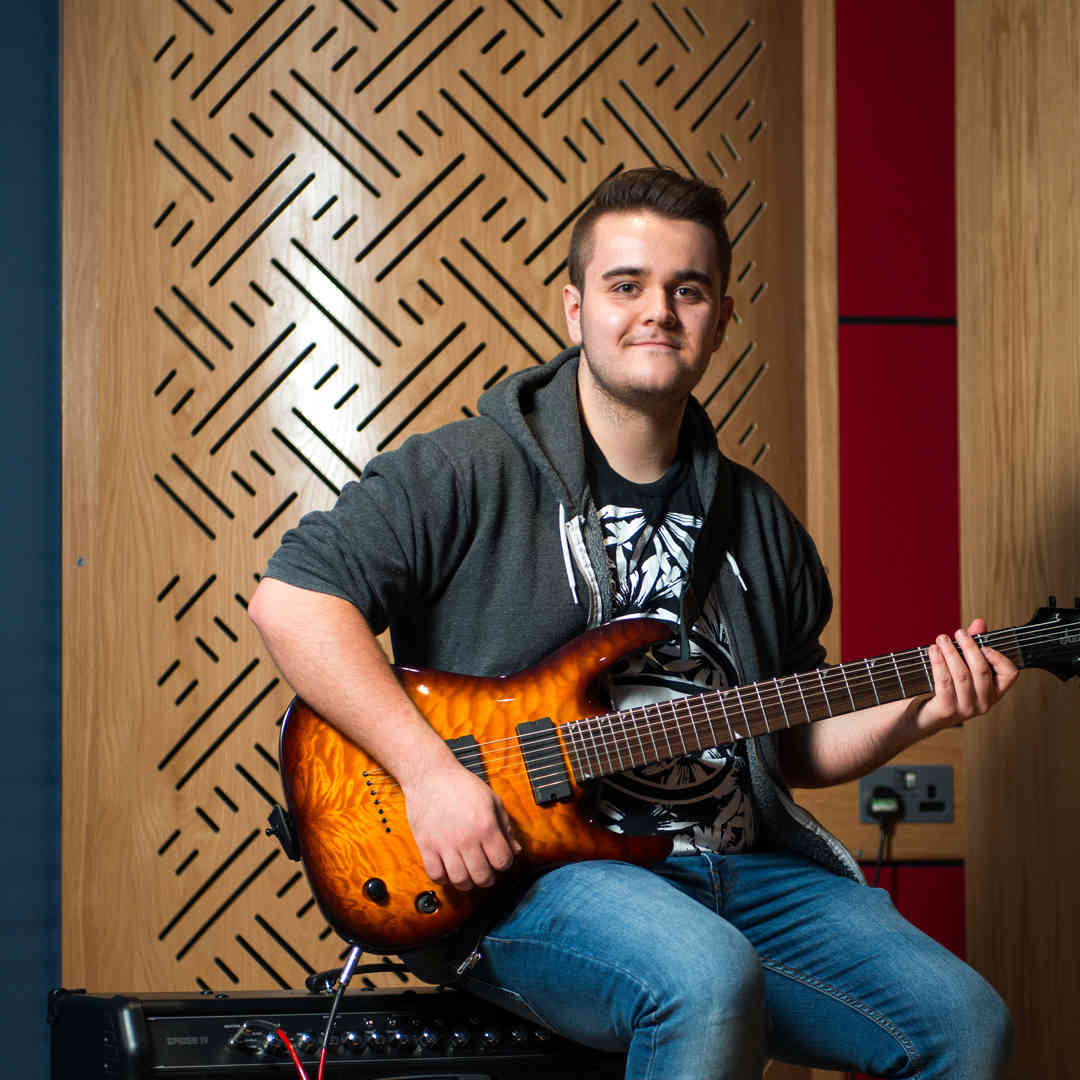 Hull Music student, Alexander Duffell, sits smiling on an amp while holiding a guitar in a recording studio.