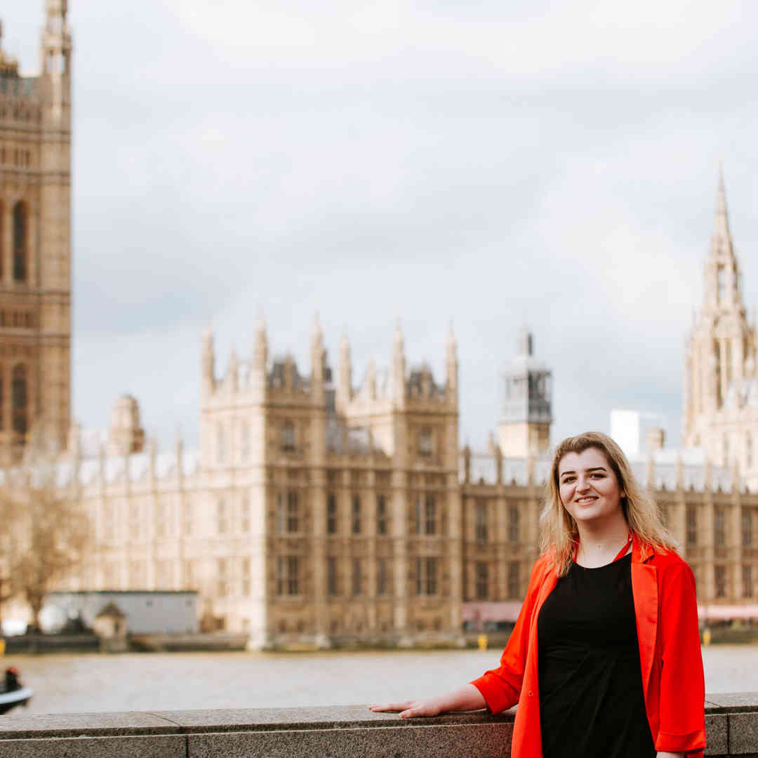 Politics student, Annemarie Wattley, stands smiling by the River Thames with the Palace of Westminster in the distance.