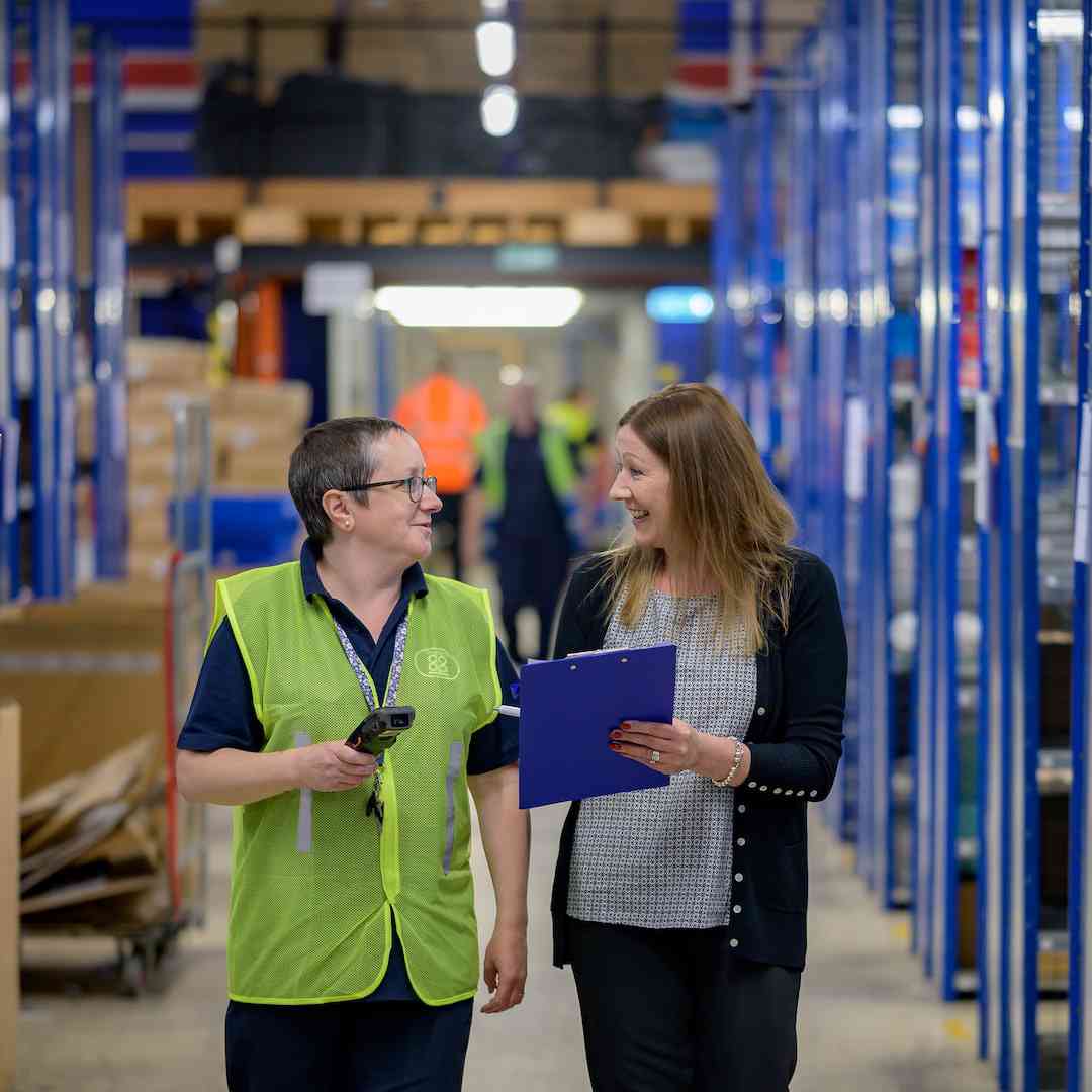A factory workers in a hi vis vest talks to a colleague holding a clipboard