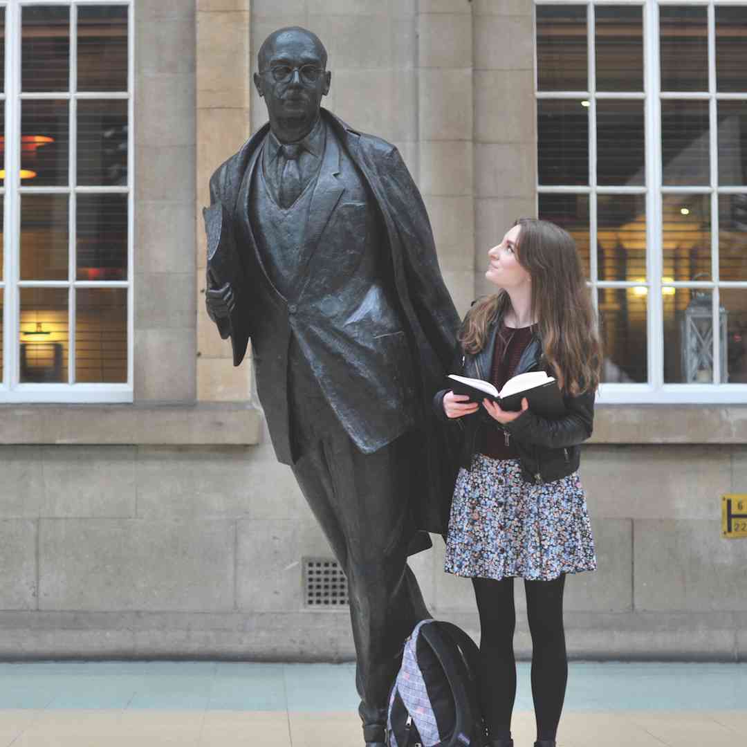 A student holding an open book stands next to a bronze statue of Philip Larkin in Hull Interchange
