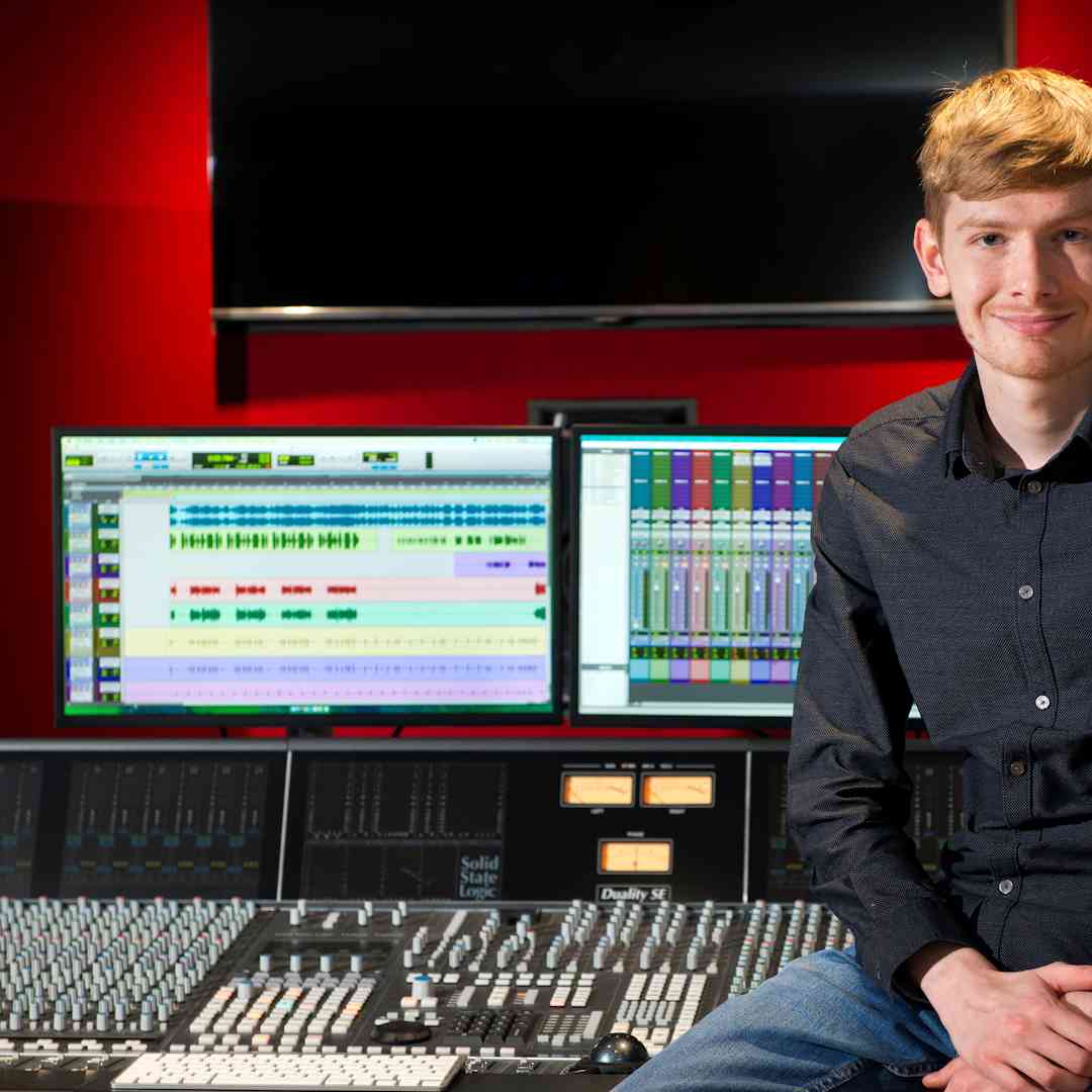 Hull Music student, Tom Wardman, sits smiling with a recording studio mixing desk and screens behind them.