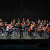 An orchestral music ensemble, including strings, brass and percussion, performing on a stage.