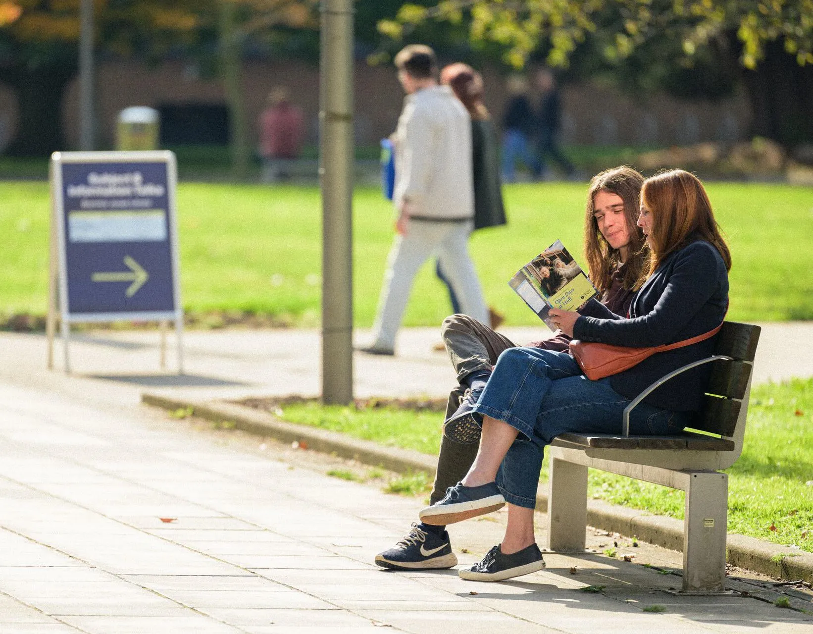 A student and his mum sit on a bench on campus on a sunny day looking at an Open Day guide