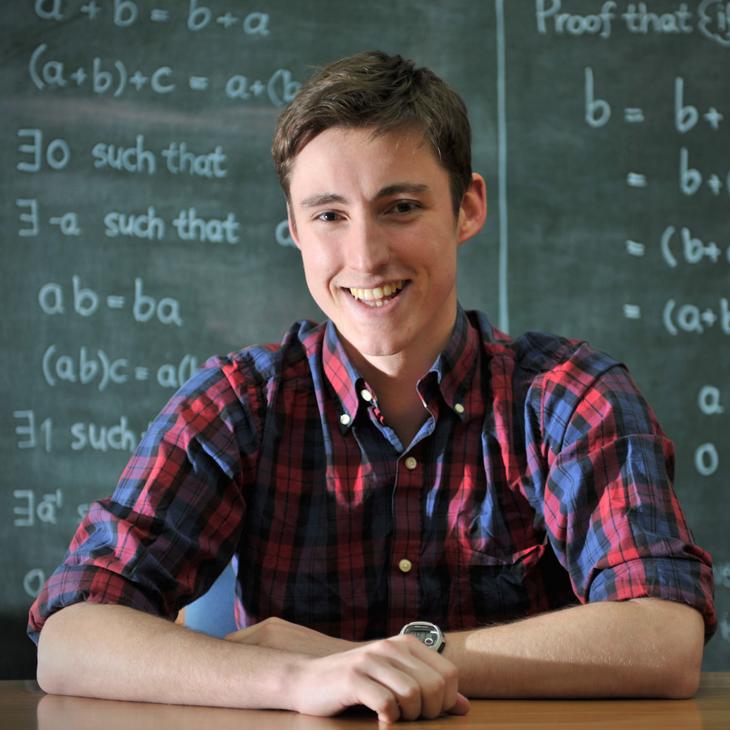 A maths student sitting in front of a chalkboard that has equations written on it