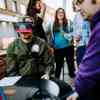 A group of students gather round an outdoor table with one wearing a virtual reality headset
