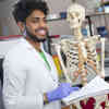 A biomedical engineering student in a lab coat and gloves, standing next to an anatomical model of a human skeleton.