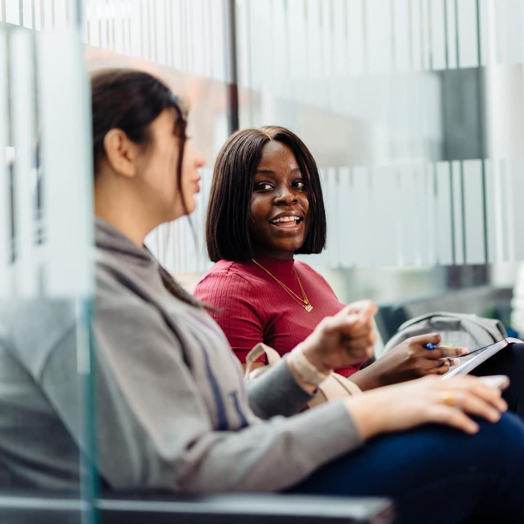 Two students sit chatting in a social space inside the Nidd building