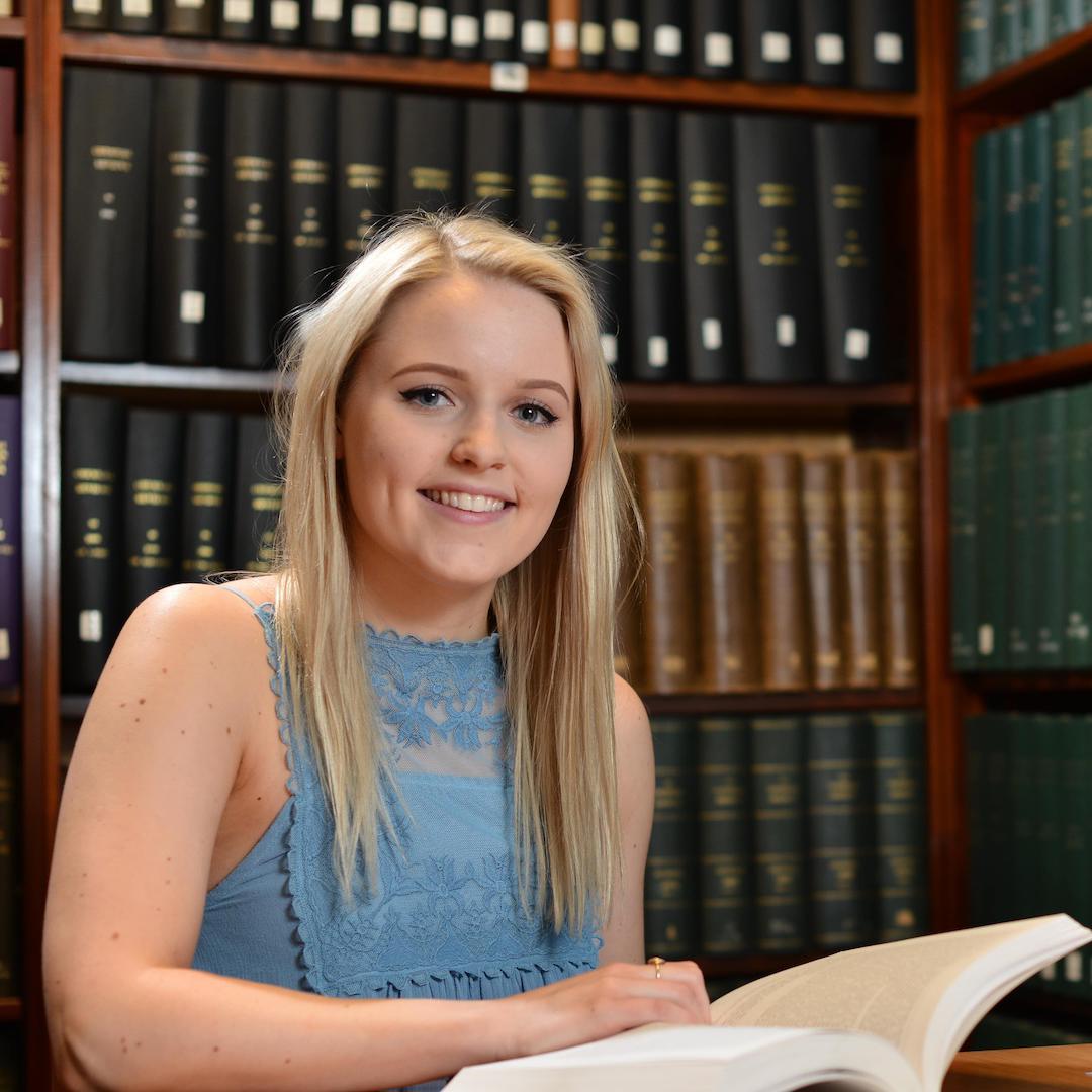 A history student sitting in a library reading a book with historical novels on the shelves behind her