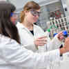 Two female students in white lab coats and protective goggles working with samples in a lab