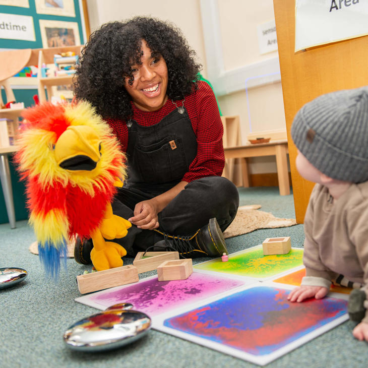 A teacher smiles at a baby while playing with a parrot puppet in a classroom