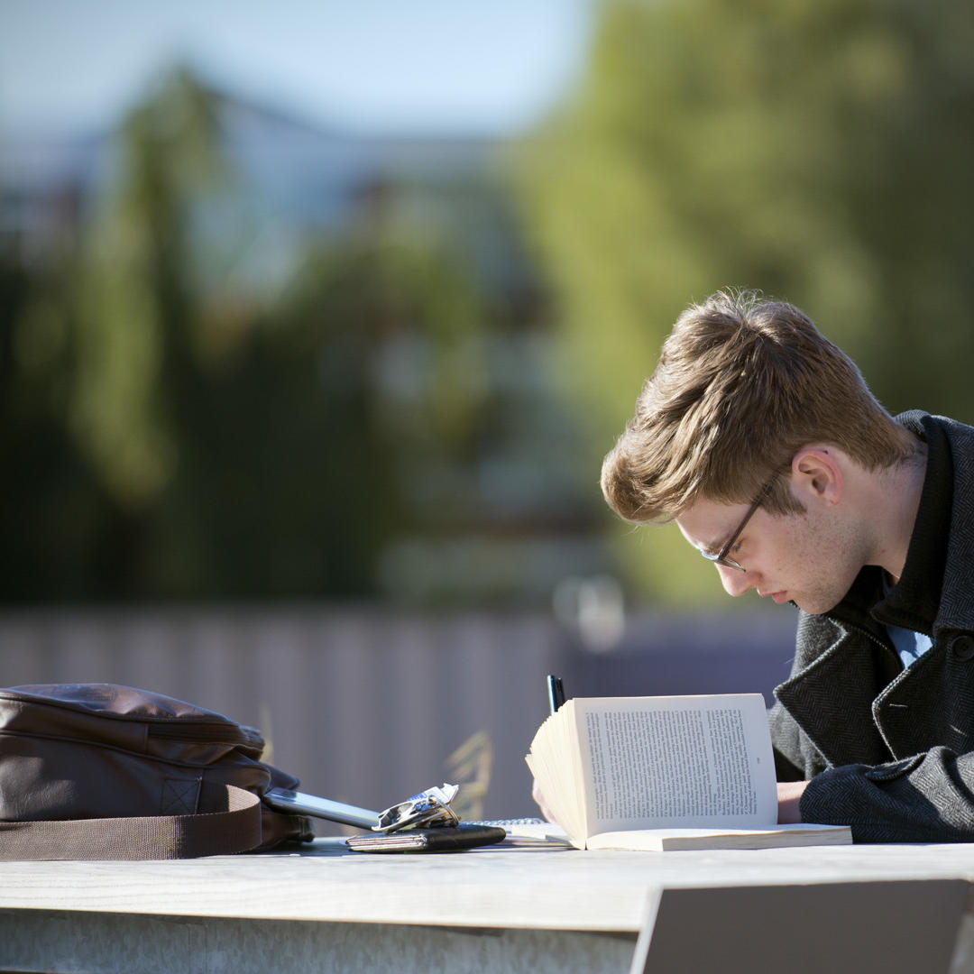 A male student writing on a notepad while working on a picnic bench with an open book beside him