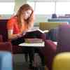 A student reads a book, surrounded by. colourful sofas in a sunny library