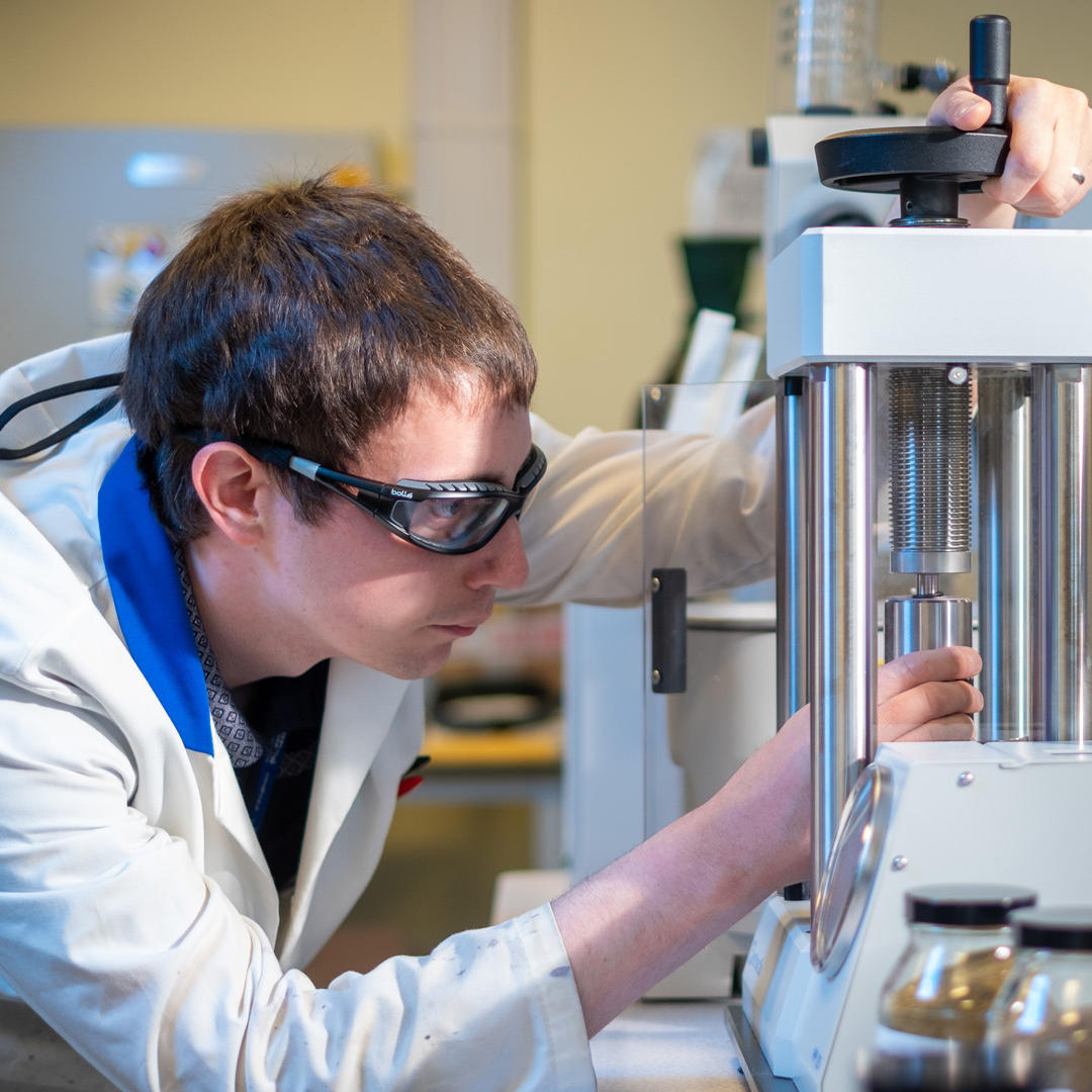 A lecturer wearing a white coat and protective goggles working on a piece of equipment in a lab 