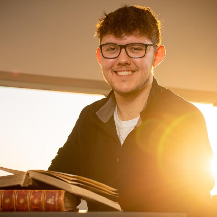 Hull History student, Dane Mellows, sits smiling with an open book and the sun resplendent behind him.