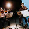 Two members of a string quartet sit playing violin on stage with sheet music on stands in front of them.