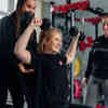 A female student lifting weights in a gym with another student standing behind her assisting