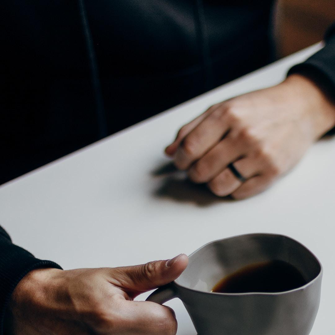 A close up of two people's hands holding mugs of coffee while sat at a table.