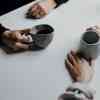 A close up of two people's hands holding mugs of coffee while sat at a table.