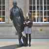 A student holding an open book stands next to a bronze statue of Philip Larkin in Hull Interchange