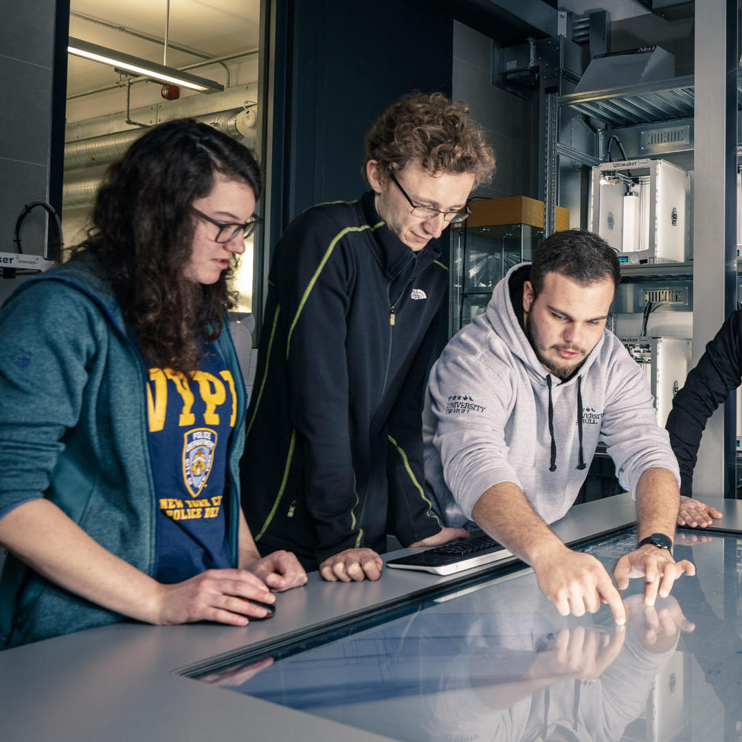 Three Hull students and one lecturer use the giant touch screen in the FabLab, with 3D printers in the background.