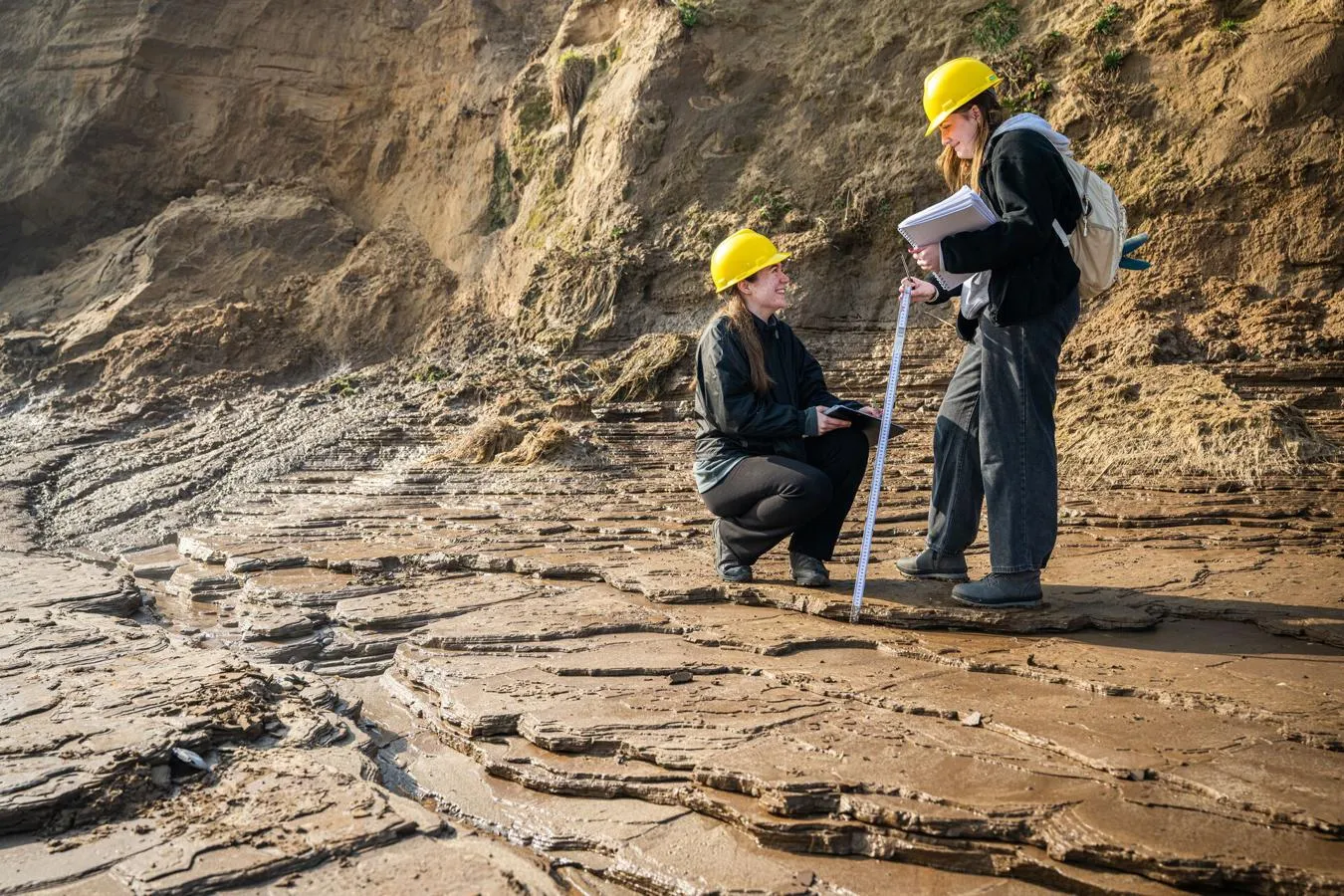 Two students in hard hats holding measuring apparatus, looking at different types of rock and sediment on a field trip