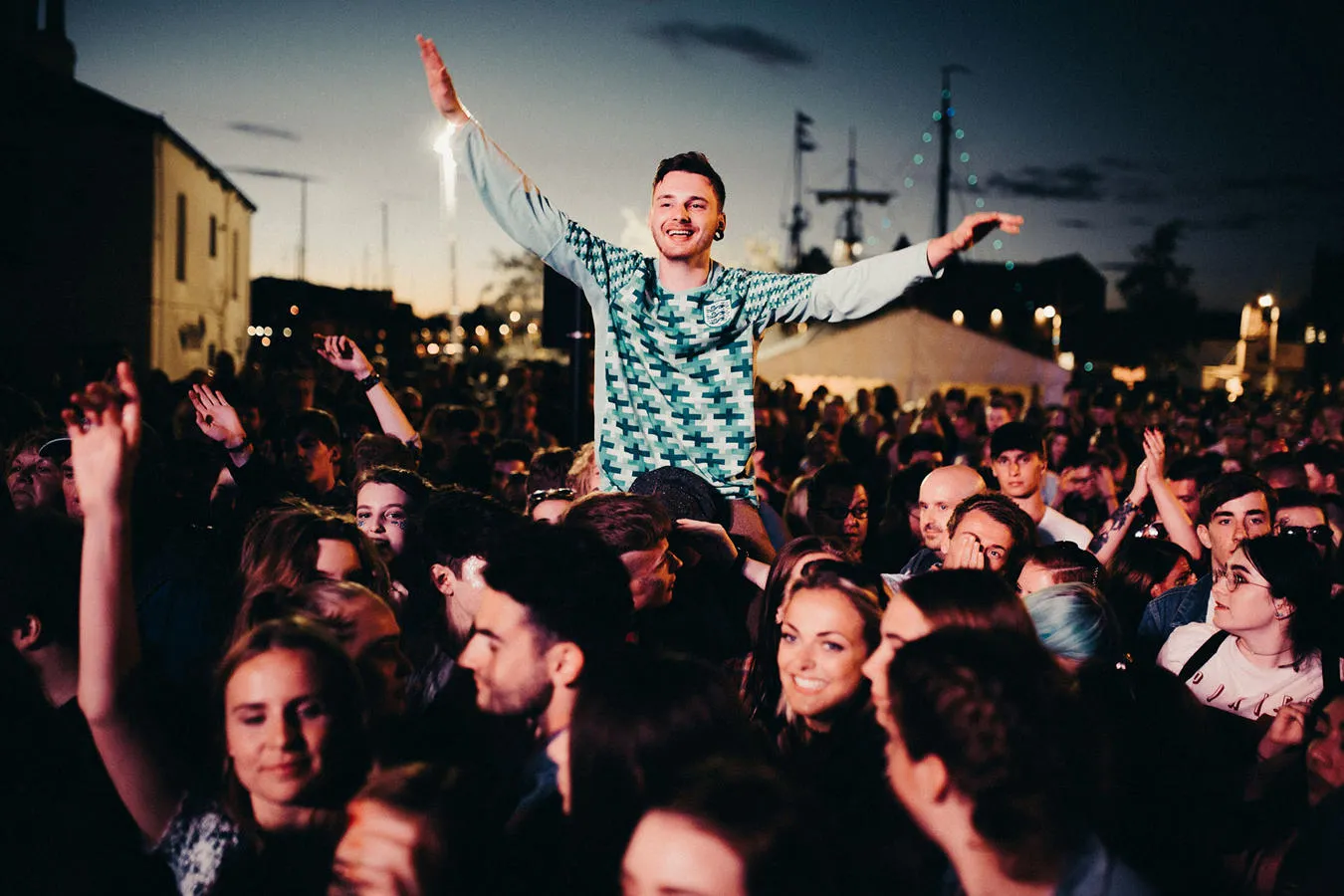 Festival crowd with one reveller sat on someone's shoulders enjoying a band at night