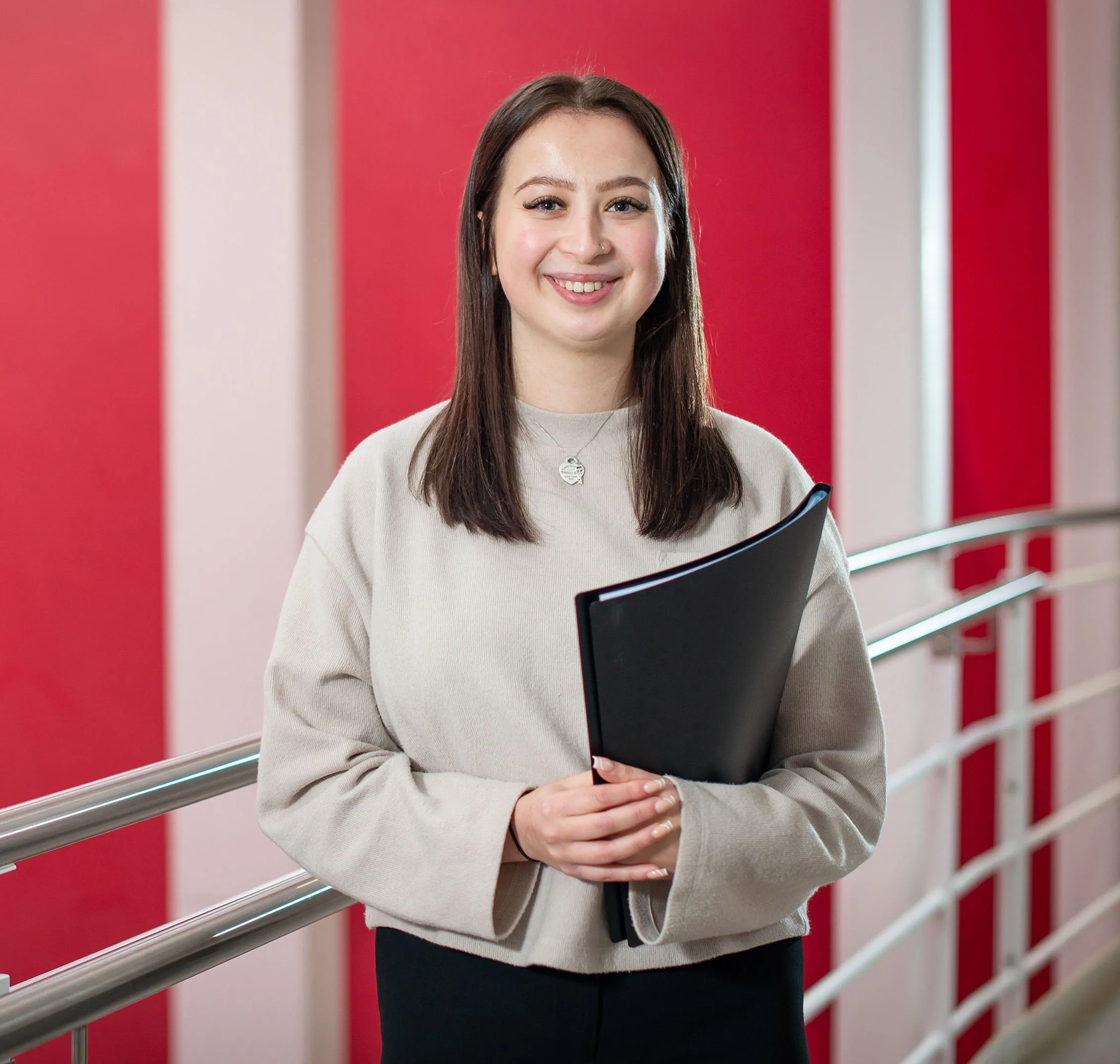 Smiling student holding a folder stands by a railing against red and white background