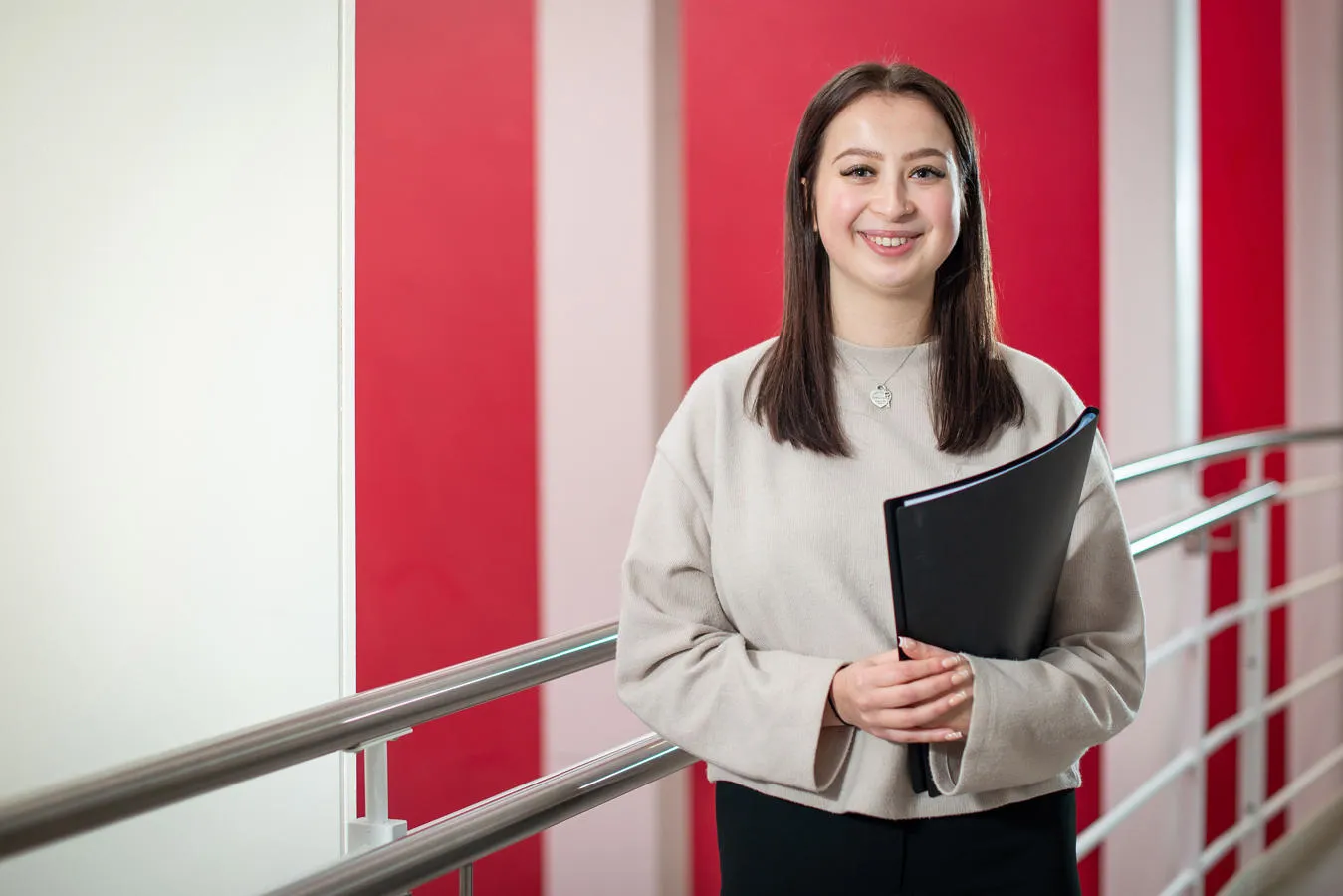 Smiling student holding a folder stands by a railing against red and white background