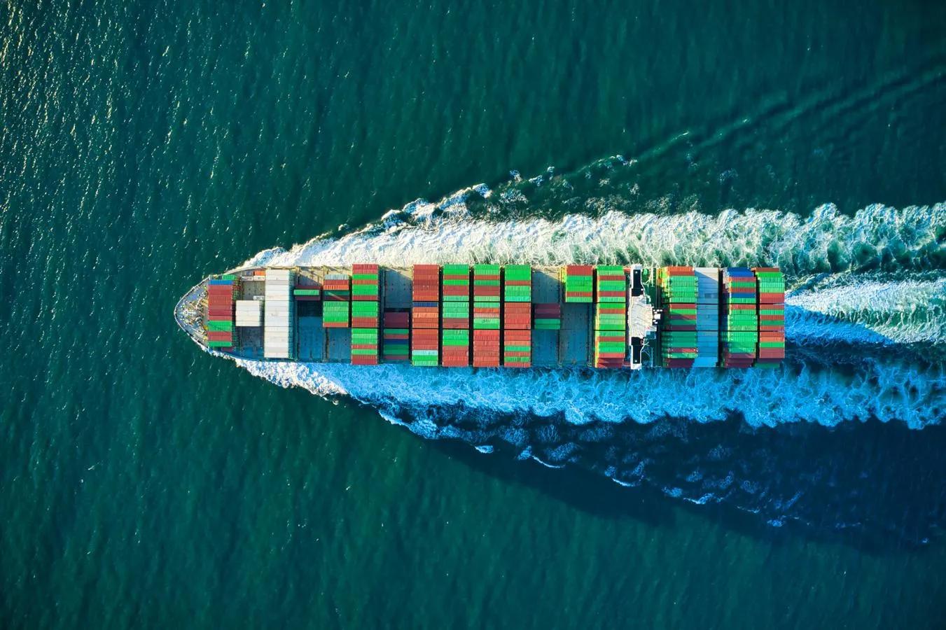 Aerial view of a boat loaded with colourful shipping containers, travelling at speed through blue waters