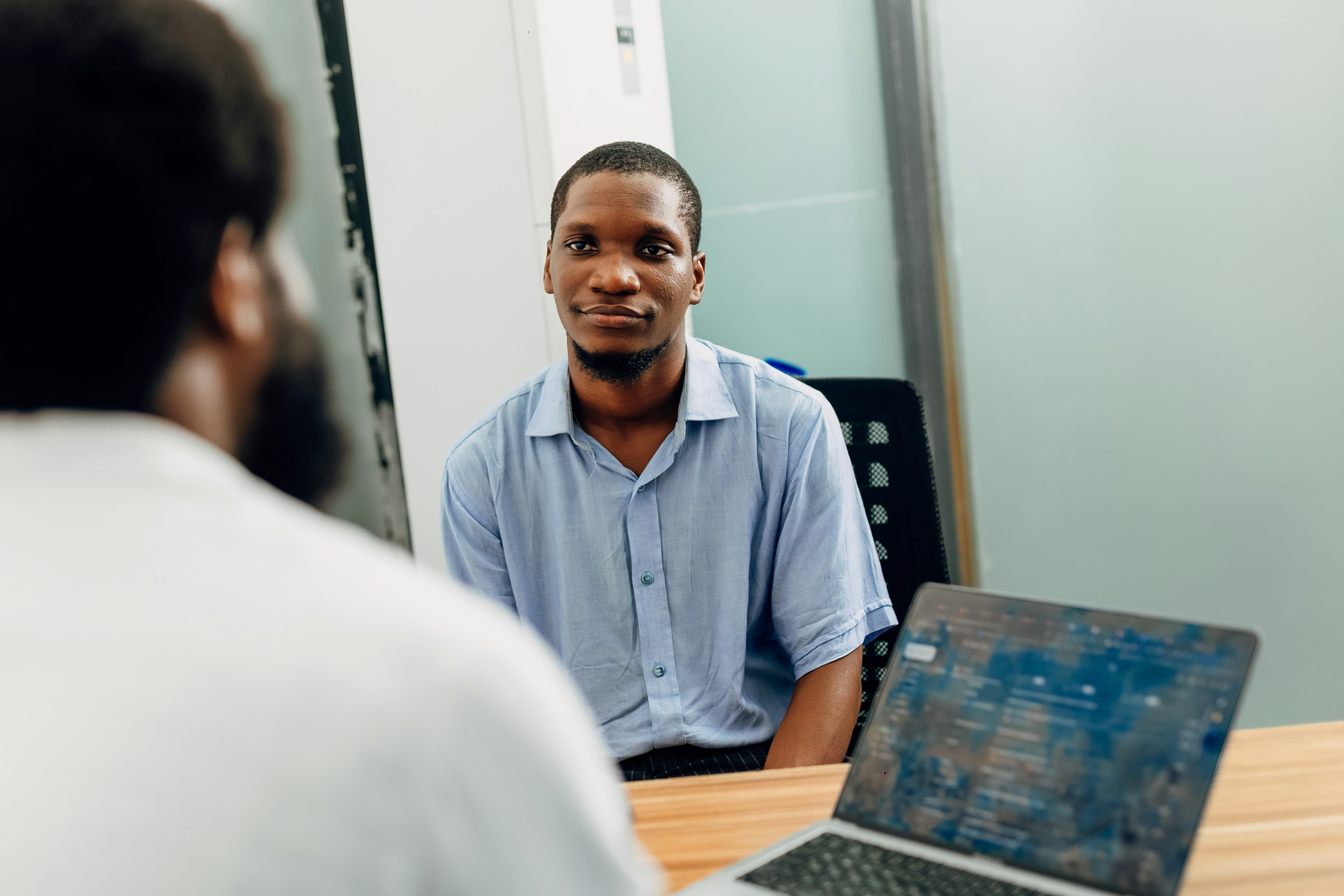 Two people having a conversation in an office setting, with a laptop on the table between them.