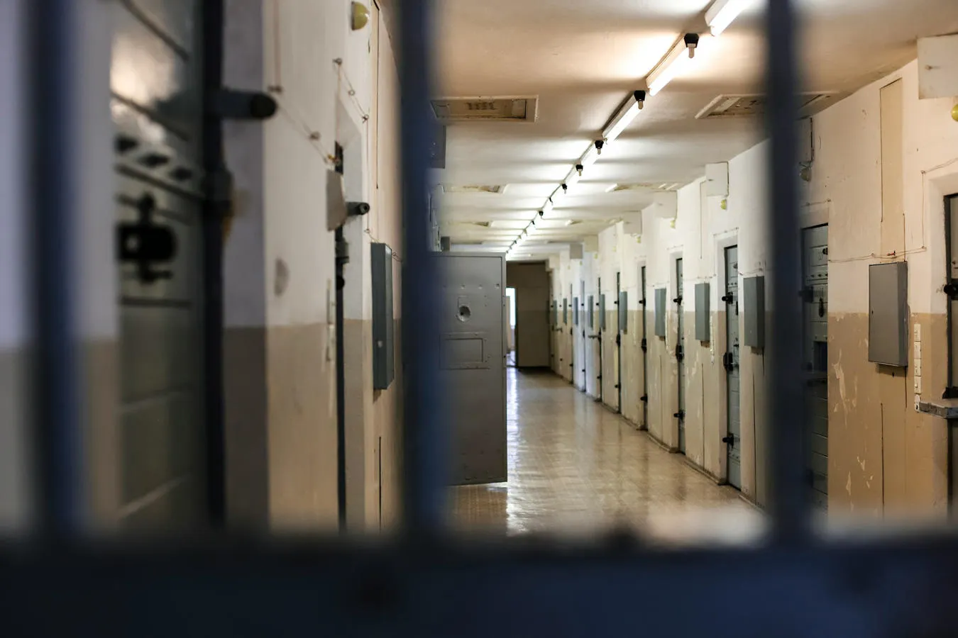 A line of closed doors in a prison interior is visible through metal bars