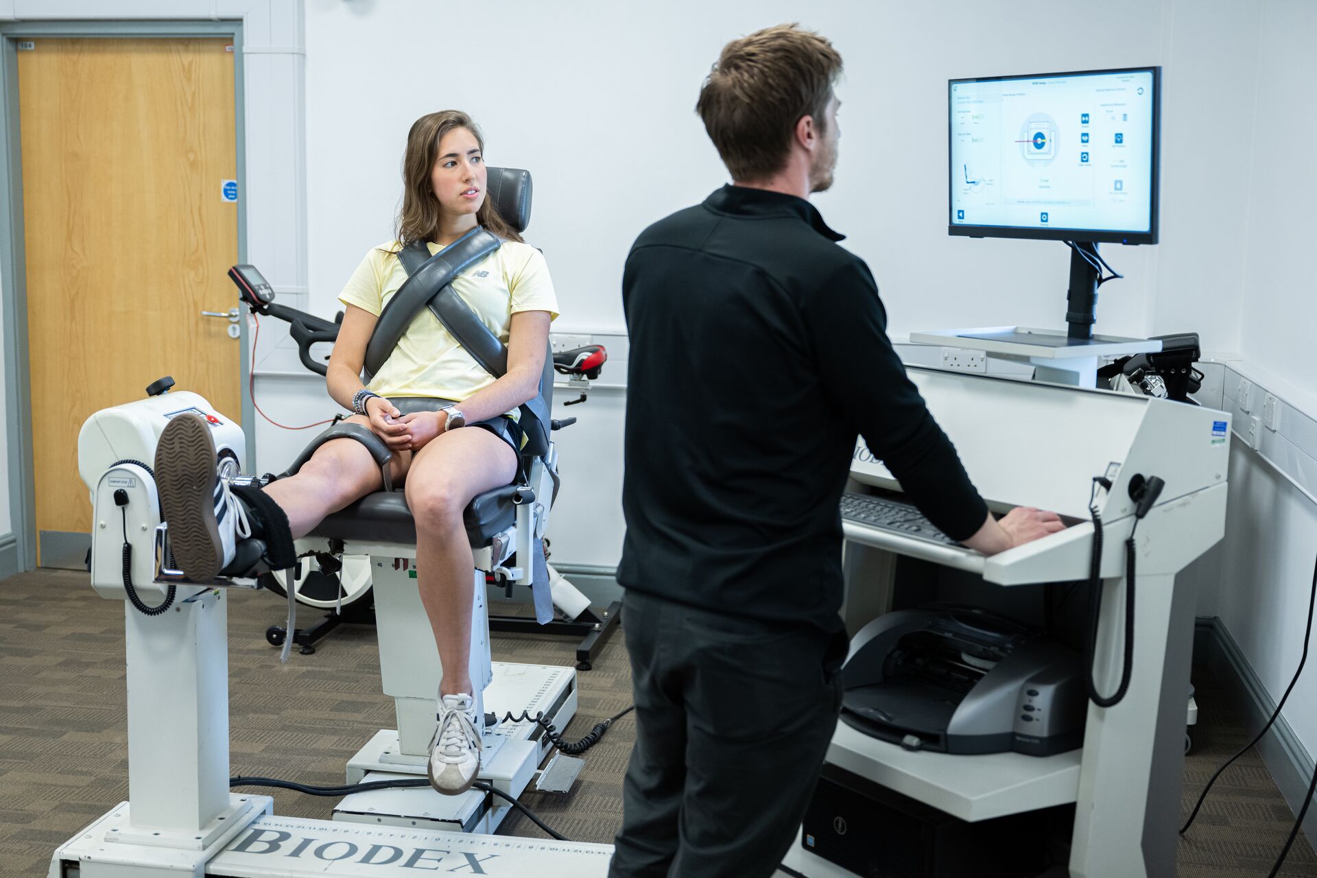 A person undergoes a leg strength test on specialised equipment while a technician monitors results on a computer.