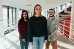 Three students stand smiling confidently on the top floor of the Business School