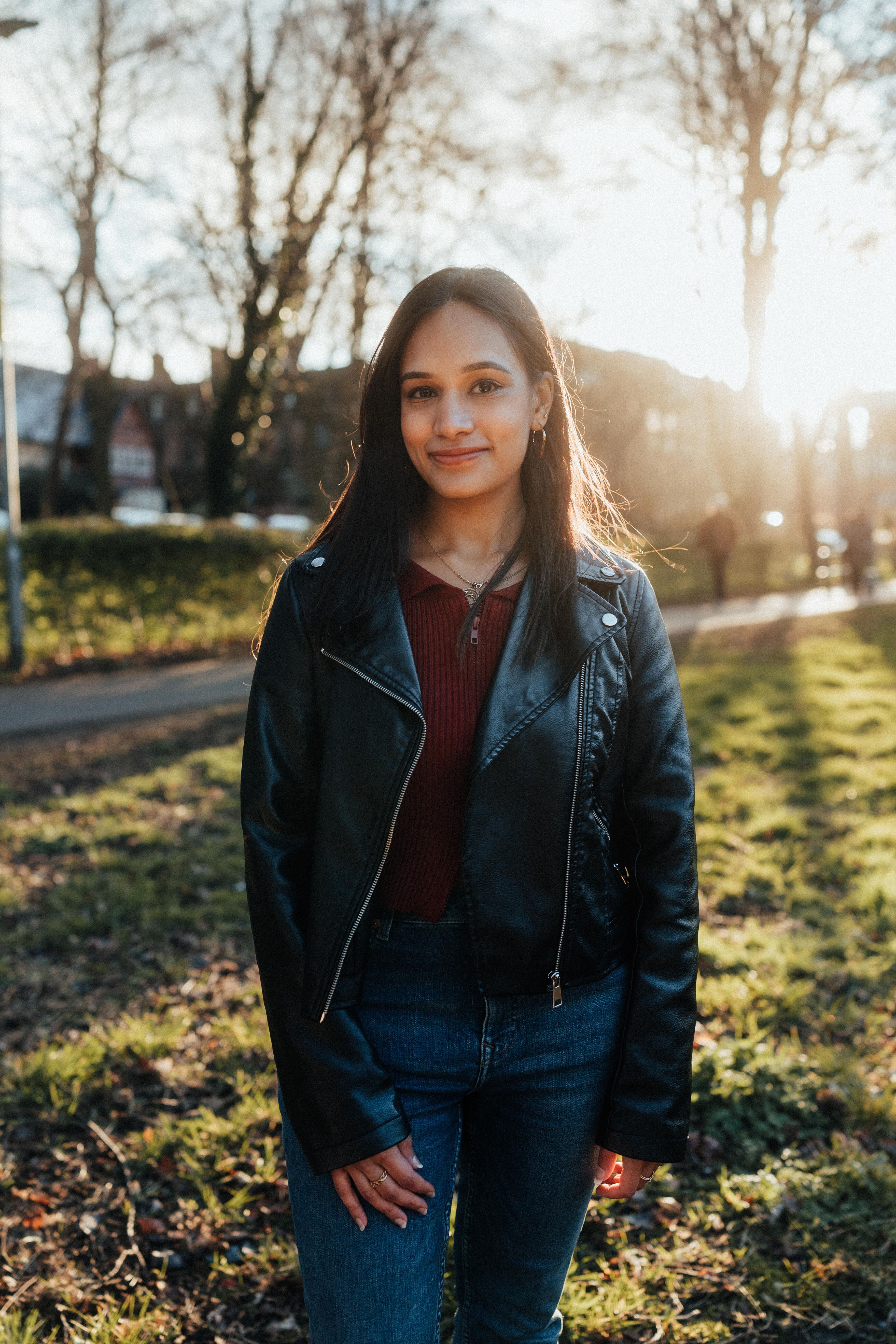 Student Aditi stands smiling in a leather jacket with the sun setting behind trees in a wooded area on campus