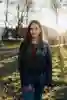 Student Aditi stands smiling in a leather jacket with the sun setting behind trees in a wooded area on campus