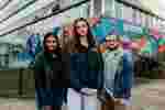 Three students stand smiling in front of a colourful spray-painted mural on the Wilberforce building