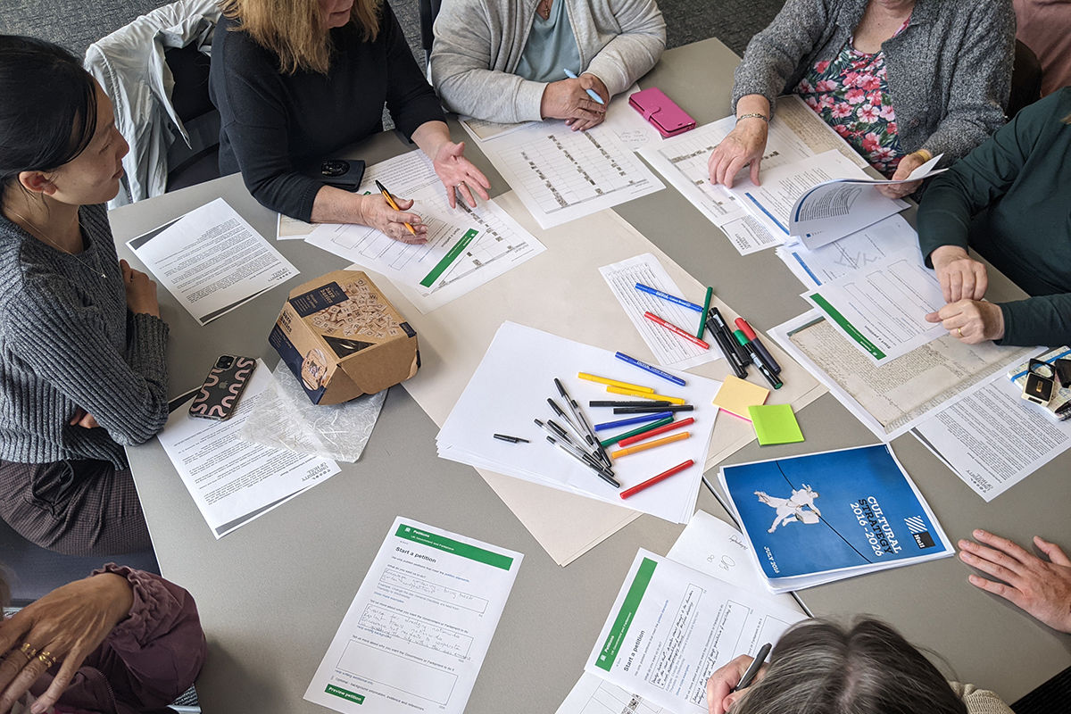 Workshop participants around a table filled with printed sheets, pens and paper