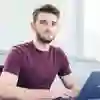 A young male student sat at a desk with a laptop looking directly the camera