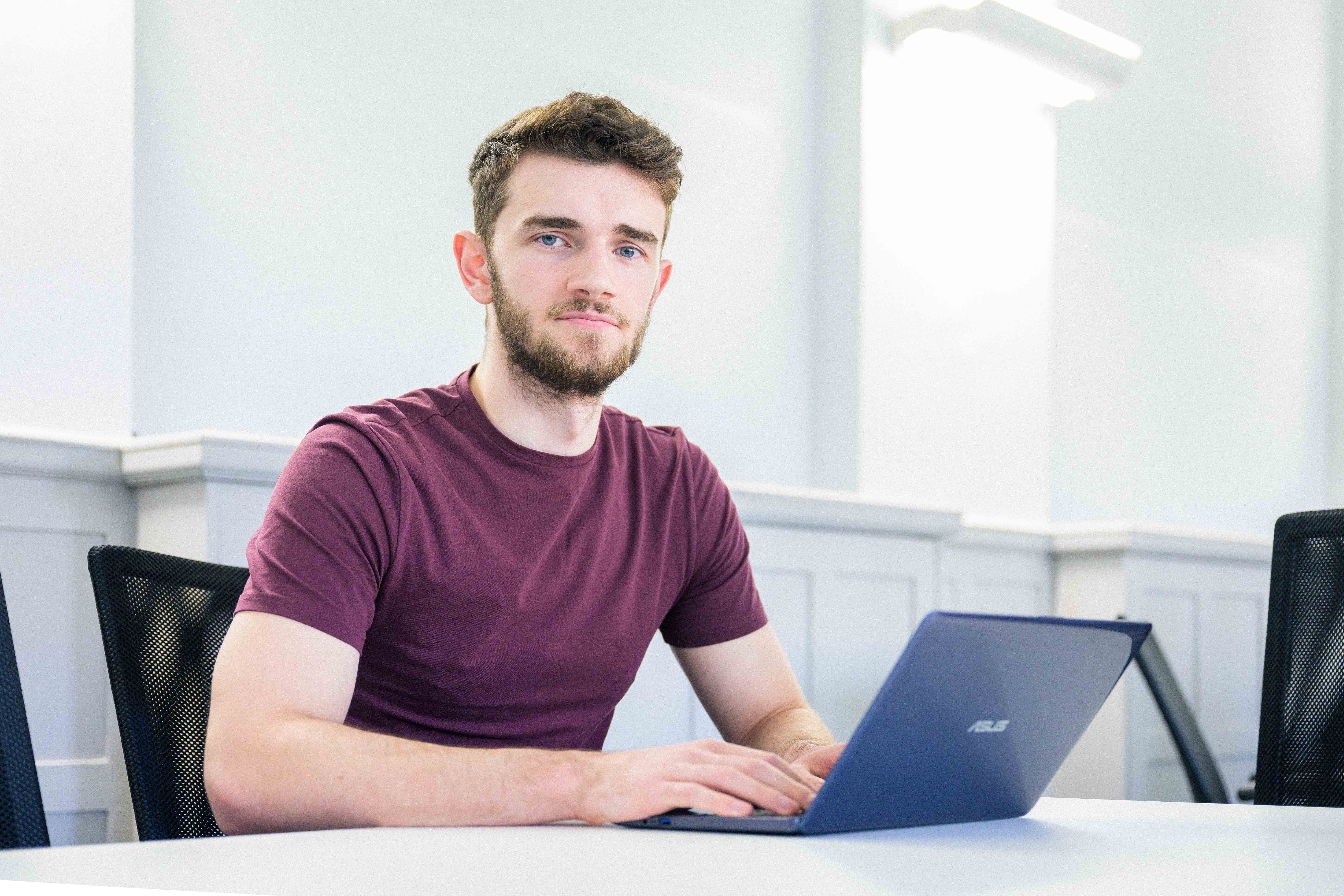 A young male student sat at a desk with a laptop looking directly the camera