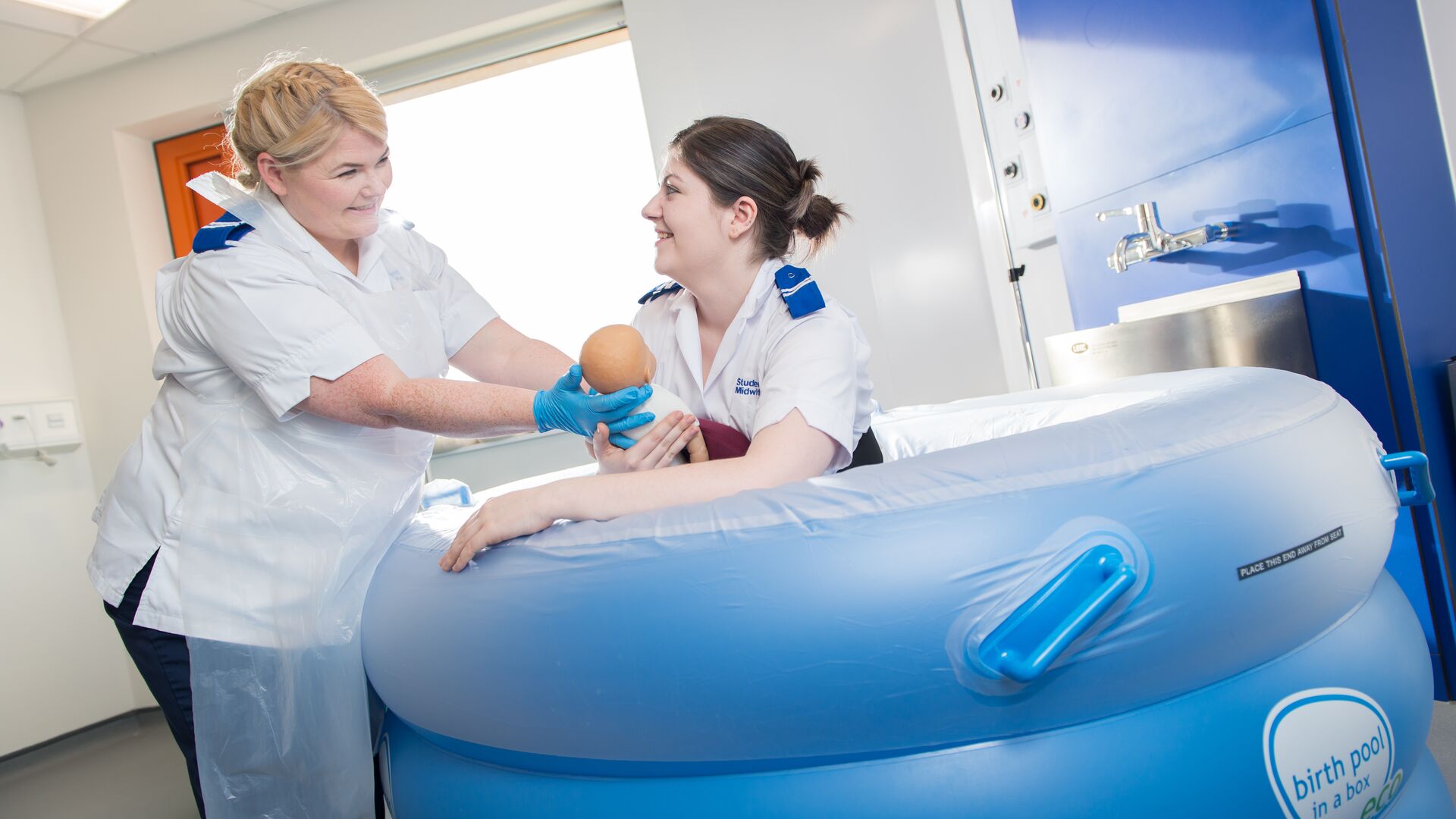 Two student midwives in white uniforms train with a dummy baby in a birthing pool