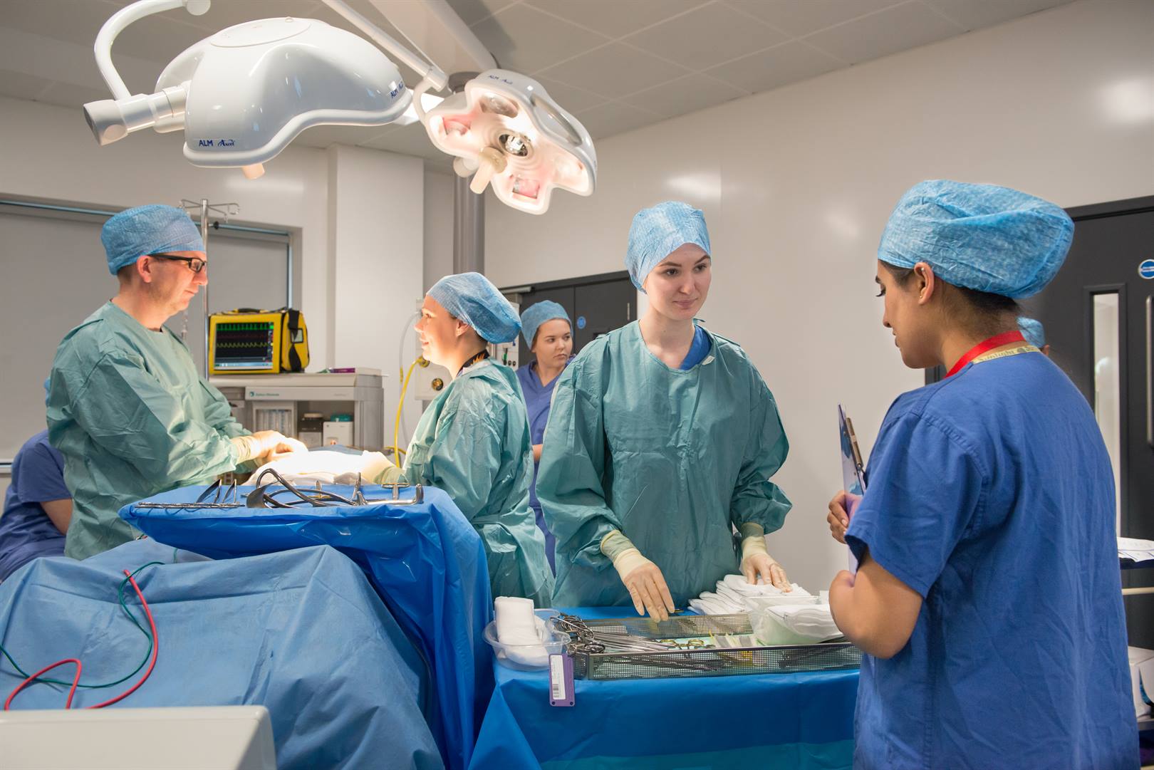 Female students wearing scrubs in a mock operating theatre with medical devices in front of them