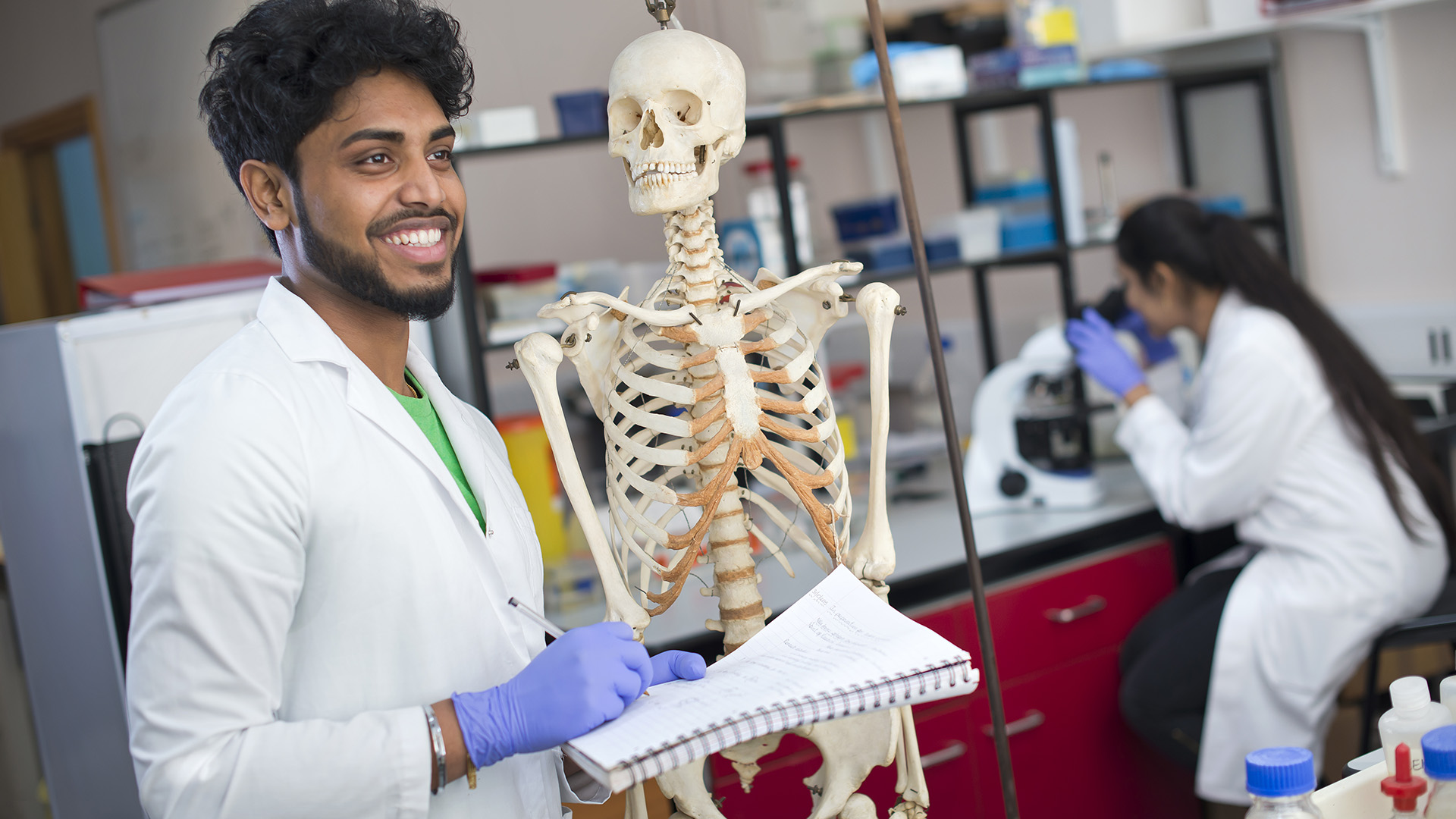 A happy male student in a white lab coat holds a notepad and pen taking measurements of a skeleton.
