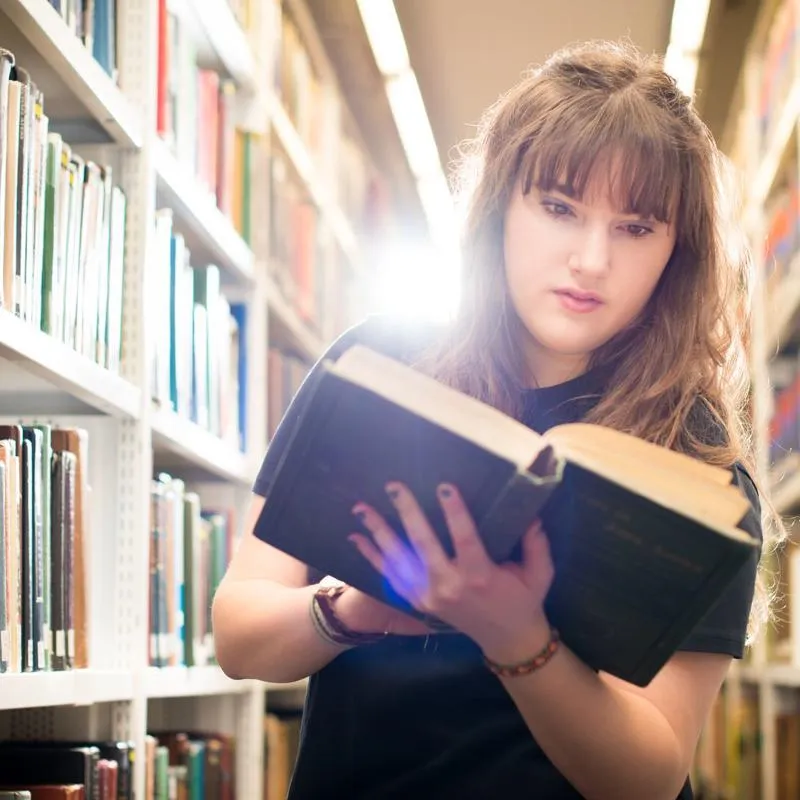 A Hull Philosophy student with an open book while browsing the bookshelves of the library.