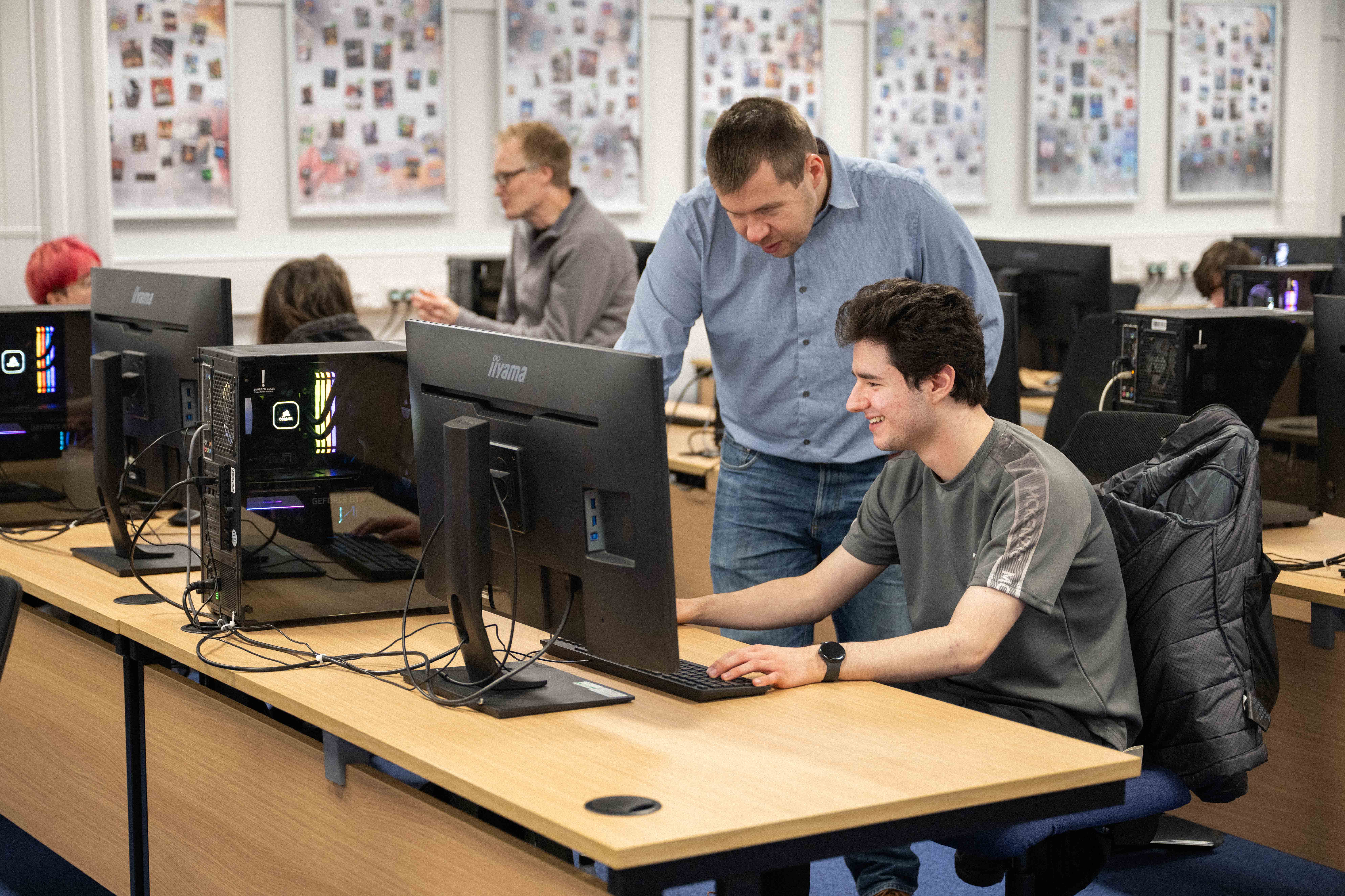 Computer science student sat working on a computer while a lecturer looks over his shoulder at his screen