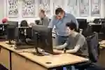 Computer science student sat working on a computer while a lecturer looks over his shoulder at his screen