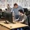 Computer science student sat working on a computer while a lecturer looks over his shoulder at his screen